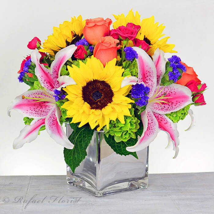 An Arrangement Of Sunflowers Stargazer Lilies Orange Roses In Mirror Vase